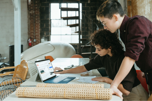 Two people collaborating at a table with a laptop, reviewing designs and plans in a modern industrial-style workspace.