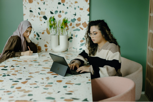 Two women working at table; one using a laptop, the other writing, in a stylish room with green walls and plants.